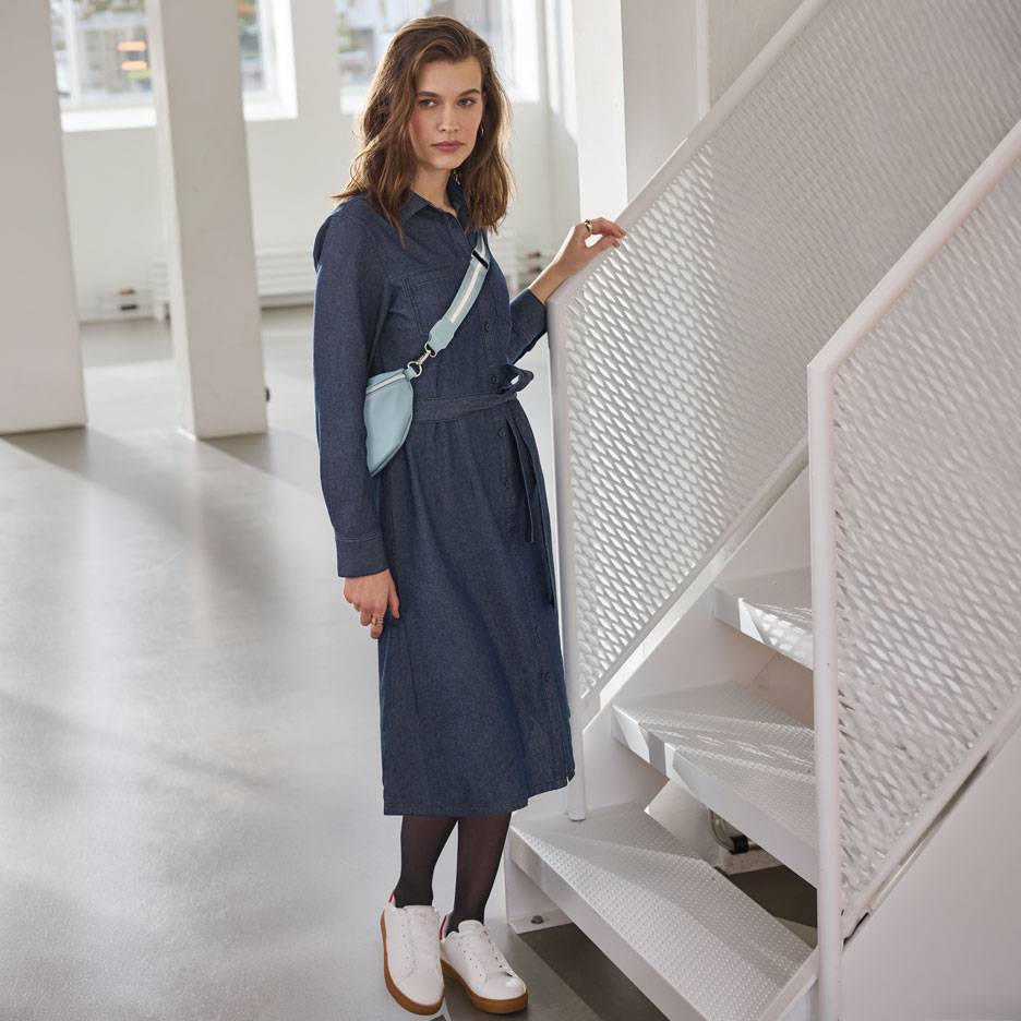 Nainen farkkupaitamekossa, mustissa sukkahousuissa ja valkoisissa tennareissa, vaaleansinisellä vyölaukulla.