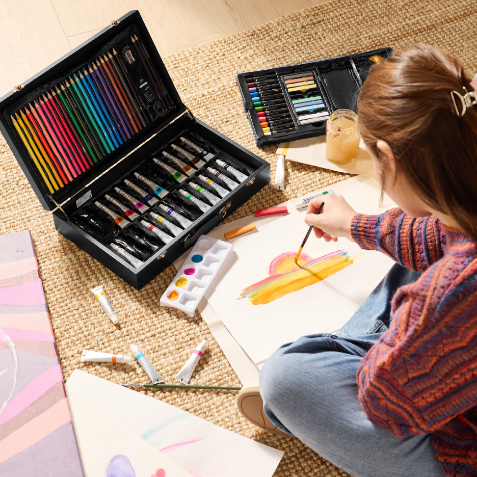 Nainen maalaa akryyliväreillä ja siveltimellä, taustalla taidetarvikesetti.