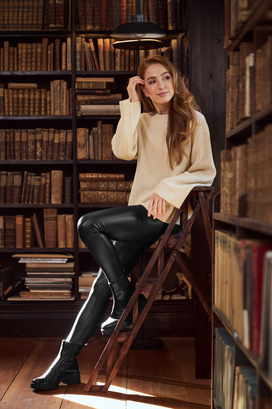 Nainen beigessä neuleessa ja mustissa nahkahousuissa kirjahyllyjen edessä.