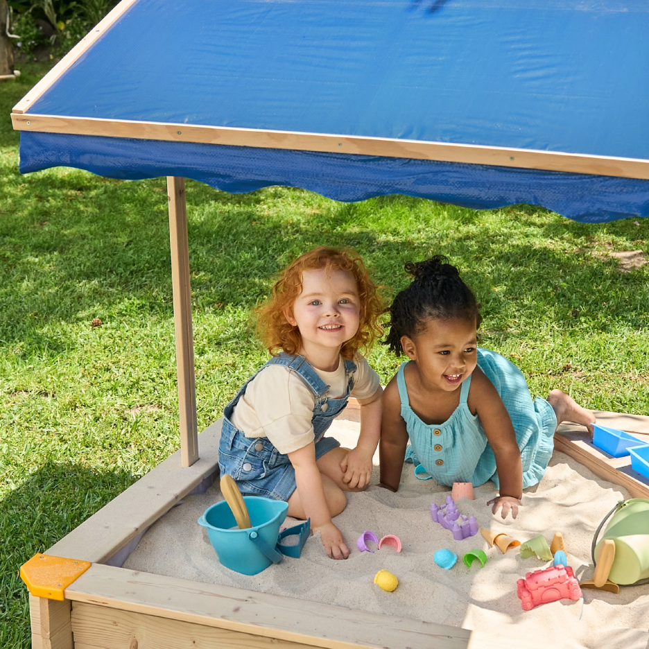 Kaksi lasta leikkii hiekkalaatikossa, jossa on sininen aurinkosuoja ja hiekkaleluja.