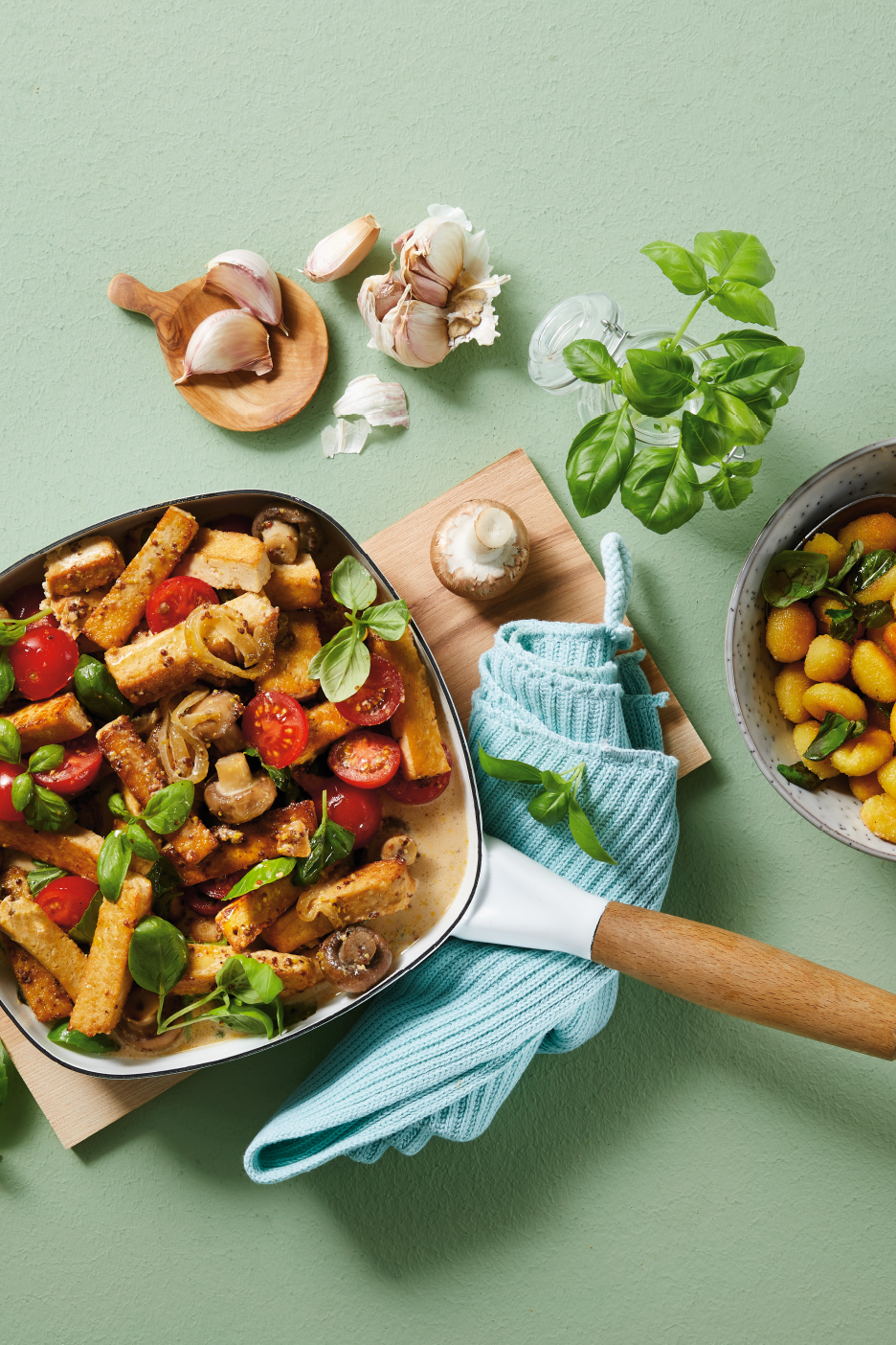 Paistinpannu täynnä tofua, tomaatteja ja sieniä, valkosipulia ja basilikaa vihreällä taustalla.